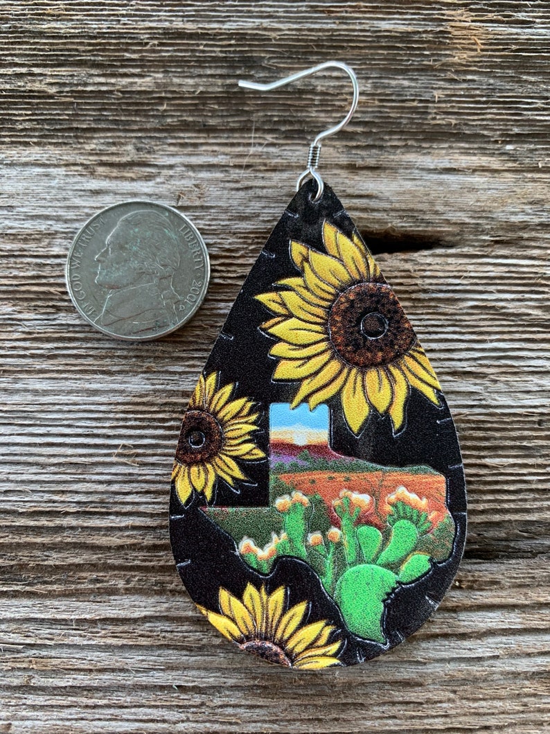 May include: A pair of dangle earrings featuring a black leather teardrop shape with a Texas outline and cactus design. The earrings are decorated with a sunflower pattern.