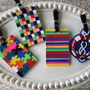 May include: Four colourful luggage tags with different designs. One tag features a jigsaw pattern with red, orange, yellow, green, blue, and black pieces. Another tag has a chequered pattern with pink, red, green, yellow, blue, and white squares. A third tag has a striped pattern with red, orange, yellow, green, blue, and pink stripes. The final tag has a blue anchor with a white rope and a red lifebuoy.