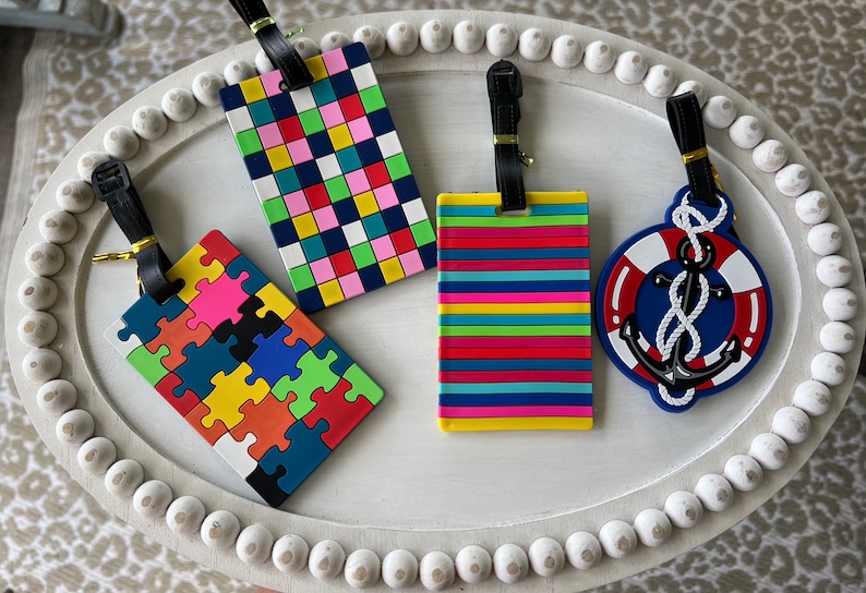 May include: Four colourful luggage tags with different designs. One tag features a jigsaw pattern with red, orange, yellow, green, blue, and black pieces. Another tag has a chequered pattern with pink, red, green, yellow, blue, and white squares. A third tag has a striped pattern with red, orange, yellow, green, blue, and pink stripes. The final tag has a blue anchor with a white rope and a red lifebuoy.