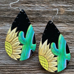 May include: A pair of teardrop-shaped earrings with a black background and a painted design of a yellow sunflower and a green cactus.