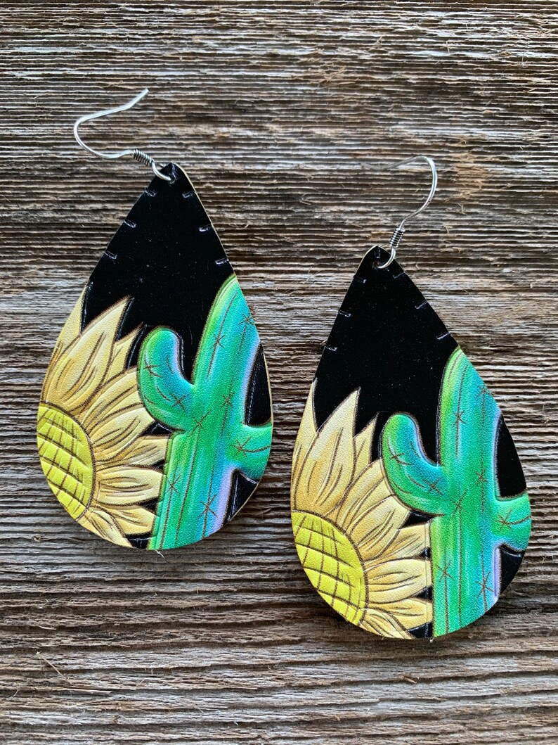 May include: A pair of teardrop-shaped earrings with a black background and a painted design of a yellow sunflower and a green cactus.