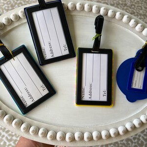 May include: Four luggage tags with black frames and white labels. The labels have the words "Name", "Address", and "Tel:" printed on them. The tags are on a white tray with a beaded edge.