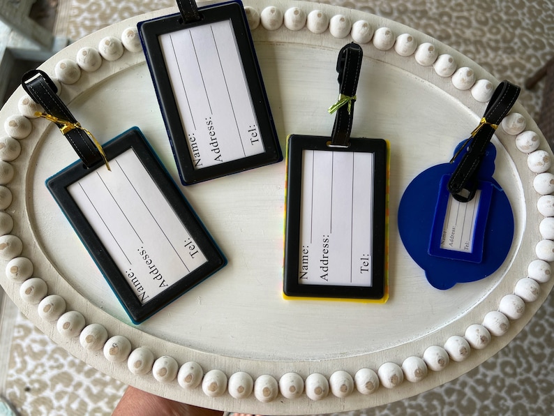 May include: Four luggage tags with black frames and white labels. The labels have the words "Name", "Address", and "Tel:" printed on them. The tags are on a white tray with a beaded edge.