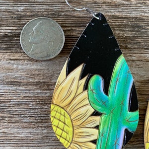 May include: A pair of dangle earrings with a black leather base and a painted design of a yellow sunflower and a green cactus. The earrings are approximately the size of a US quarter.