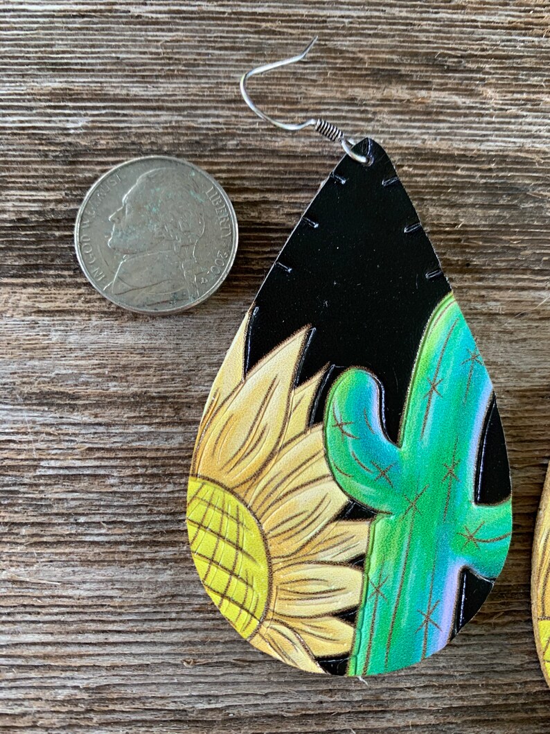 May include: A pair of dangle earrings with a black leather base and a painted design of a yellow sunflower and a green cactus. The earrings are approximately the size of a US quarter.