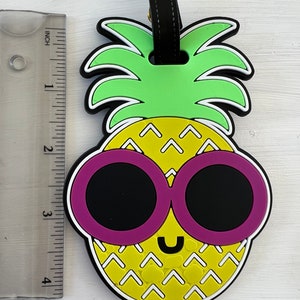 Op de afbeelding: Een gele ananas met een groene bovenkant en roze zonnebril. De ananas heeft een wit en geel patroon en een glimlachend gezicht.