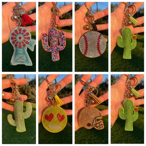 May include: A set of ten rhinestone keychains featuring various designs, including a windmill, a cactus, a baseball, a smiley face, and a football helmet. Each keychain has a gold chain and a tassel.
