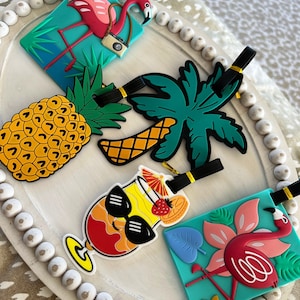 K&ouml;nnte beinhalten: Vier farbenfrohe Gep&auml;ckanh&auml;nger mit tropischen Motiven. Ein rosa Flamingo mit einer Kamera, eine Ananas, eine Palme und ein Cocktail mit Sonnenbrille.