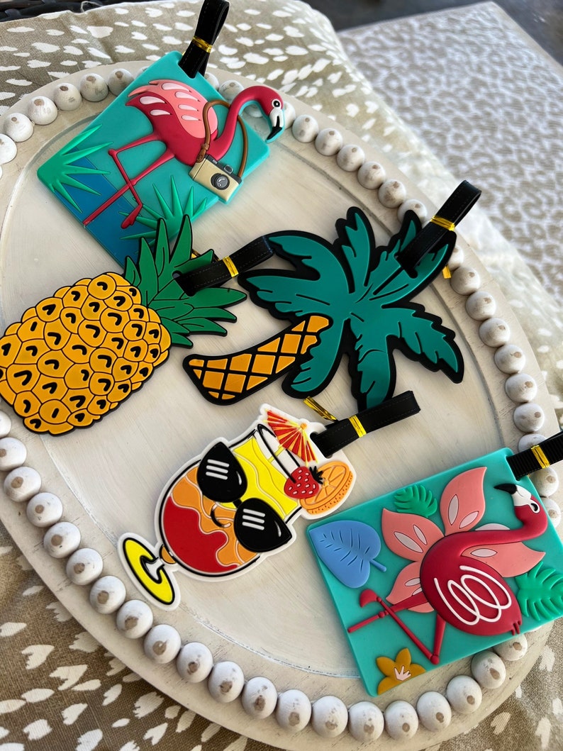 K&ouml;nnte beinhalten: Vier farbenfrohe Gep&auml;ckanh&auml;nger mit tropischen Motiven. Ein rosa Flamingo mit einer Kamera, eine Ananas, eine Palme und ein Cocktail mit Sonnenbrille.