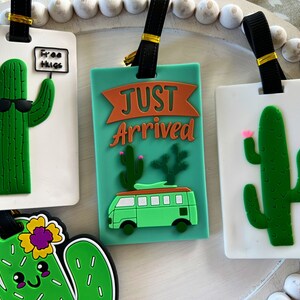 K&ouml;nnte beinhalten: Vier bunte Gep&auml;ckanh&auml;nger mit Kaktus-Motiven. Ein Anh&auml;nger sagt "Free Hugs" mit einem Kaktus, der eine Sonnenbrille tr&auml;gt. Ein anderer Anh&auml;nger sagt "Just Arrived" mit einem Kaktus und einem gr&uuml;nen Van. Die beiden anderen Anh&auml;nger zeigen einen Kaktus mit rosa Bl&uuml;ten.