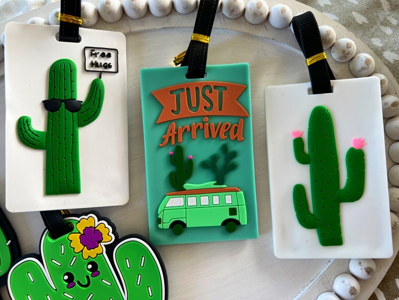 K&ouml;nnte beinhalten: Vier bunte Gep&auml;ckanh&auml;nger mit Kaktus-Motiven. Ein Anh&auml;nger sagt "Free Hugs" mit einem Kaktus, der eine Sonnenbrille tr&auml;gt. Ein anderer Anh&auml;nger sagt "Just Arrived" mit einem Kaktus und einem gr&uuml;nen Van. Die beiden anderen Anh&auml;nger zeigen einen Kaktus mit rosa Bl&uuml;ten.