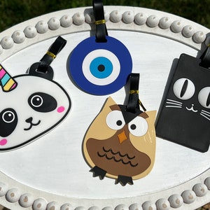 May include: Four colorful luggage tags with different designs. A black and white panda with a unicorn horn, a blue evil eye, a brown owl, and a black cat.