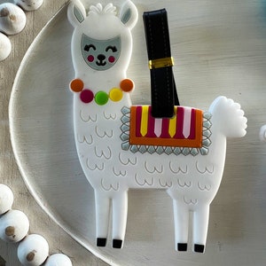 May include: White llama luggage tag with a black strap and colorful details. The llama has a smiling face and a colorful saddle.