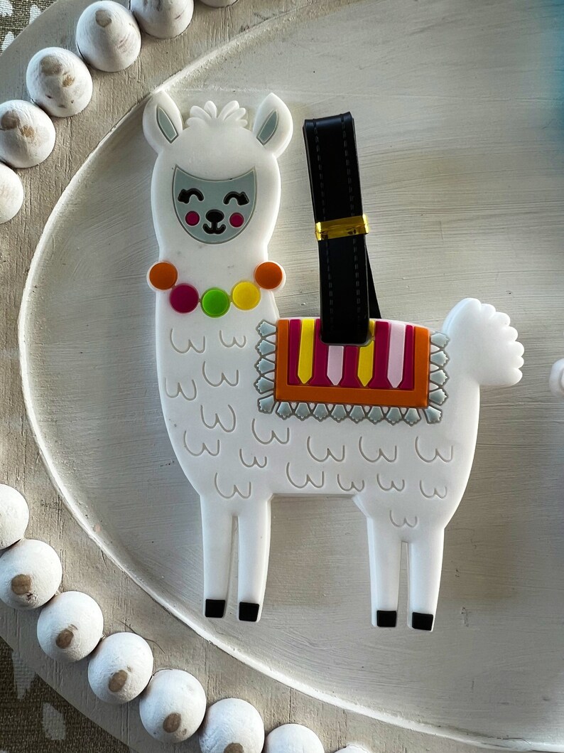 May include: White llama luggage tag with a black strap and colorful details. The llama has a smiling face and a colorful saddle.