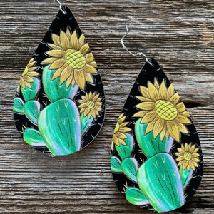 May include: A pair of dangle earrings with a black background and a green cactus and yellow sunflower design.