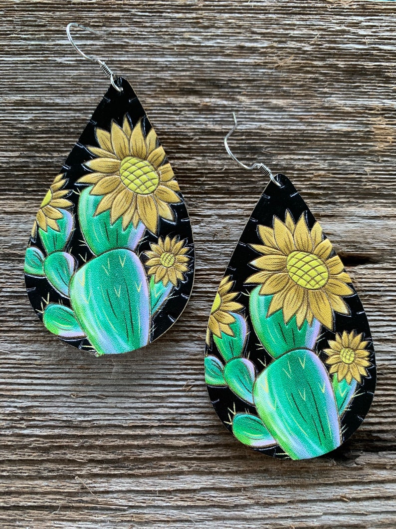May include: A pair of dangle earrings with a black background and a green cactus and yellow sunflower design.