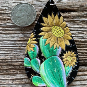 May include: A pair of dangle earrings with a black leather background and a painted design of a cactus and sunflowers. The earrings are approximately the size of a US quarter.