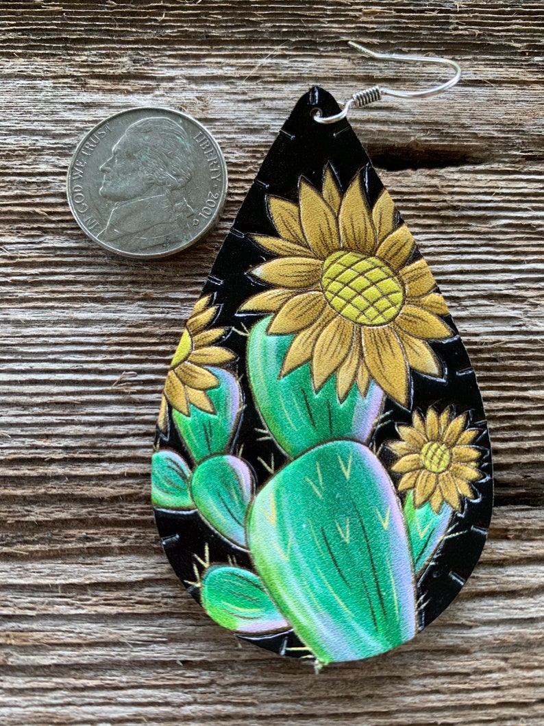 May include: A pair of dangle earrings with a black leather background and a painted design of a cactus and sunflowers. The earrings are approximately the size of a US quarter.