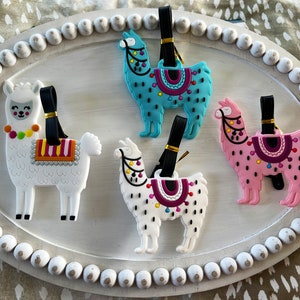 May include: Four colorful llama luggage tags with black straps. The llamas are white, pink, blue, and white with black spots. Each llama has a saddle with colorful designs.