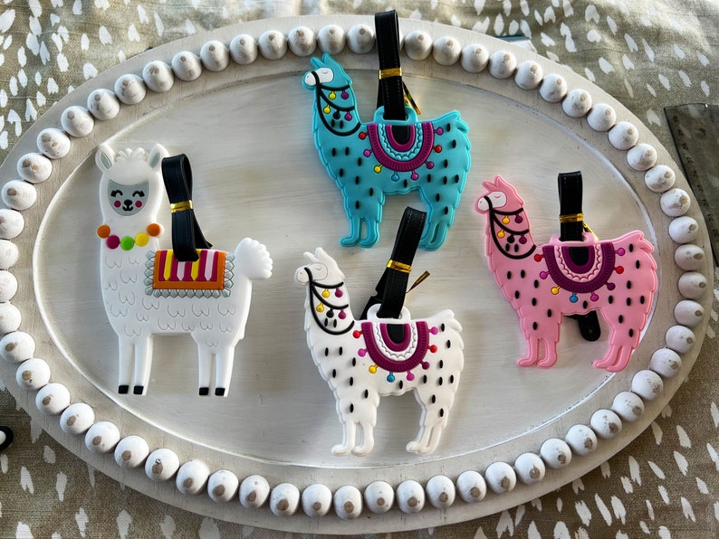 May include: Four colorful llama luggage tags with black straps. The llamas are white, pink, blue, and white with black spots. Each llama has a saddle with colorful designs.