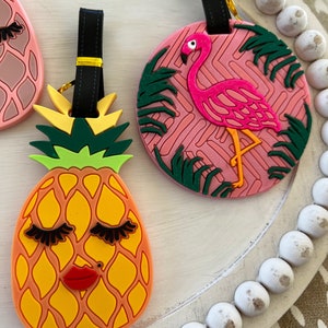 Op de afbeelding: Twee kleurrijke bagagelabeltjes met een ananas- en een flamingo-ontwerp. De ananas is geel met groene bladeren en heeft wimpers en een rode mond. De flamingo is roze met zwarte ogen en staat op &eacute;&eacute;n poot voor groene palmbladeren. Beide labeltjes hebben zwarte bandjes met gouden accenten.