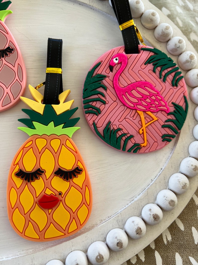 Op de afbeelding: Twee kleurrijke bagagelabeltjes met een ananas- en een flamingo-ontwerp. De ananas is geel met groene bladeren en heeft wimpers en een rode mond. De flamingo is roze met zwarte ogen en staat op &eacute;&eacute;n poot voor groene palmbladeren. Beide labeltjes hebben zwarte bandjes met gouden accenten.