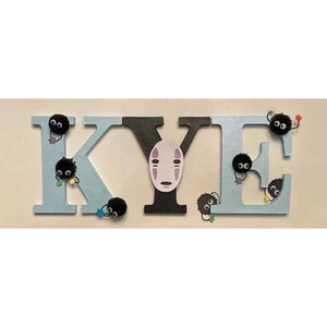 K&ouml;nnte beinhalten: Holzbuchstaben, die den Namen "KYE" in hellblau und schwarz buchstabieren. Jeder Buchstabe hat kleine schwarze, flauschige Kugeln mit Augen daran befestigt. Das "Y" hat einen wei&szlig;en No Face-Charakter aus dem Film Chihiros Reise ins Zauberland daran befestigt.