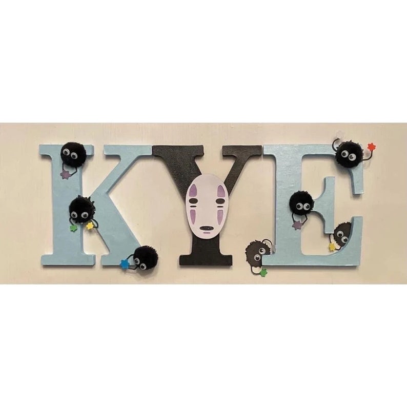K&ouml;nnte beinhalten: Holzbuchstaben, die den Namen "KYE" in hellblau und schwarz buchstabieren. Jeder Buchstabe hat kleine schwarze, flauschige Kugeln mit Augen daran befestigt. Das "Y" hat einen wei&szlig;en No Face-Charakter aus dem Film Chihiros Reise ins Zauberland daran befestigt.