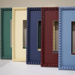 May include: A collection of colorful picture frames in shades of green, cream, navy, burgundy, and light blue. Each frame has a square opening and is decorated with a beaded border.