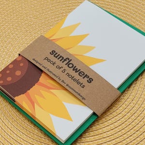 May include: A pack of five notelets featuring a sunflower illustration, secured with a brown paper band printed with the text sunflowers pack of 5 notelets designed and illustrated by Bev @oconnordesign, with green envelopes visible underneath.