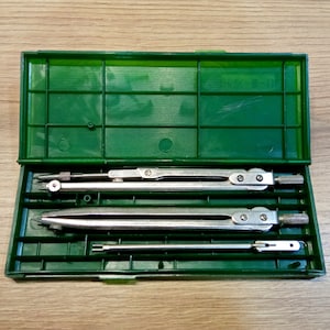 May include: A green plastic case containing three silver drafting tools. The tools include a compass, a divider, and a technical pen. The case is open, revealing the tools neatly arranged inside.
