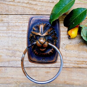 May include: A bronze lion head door knocker with a silver ring handle, fixed to a wooden surface. The knocker features a detailed lion face design. Green leaves and a small yellow pear are in the background.