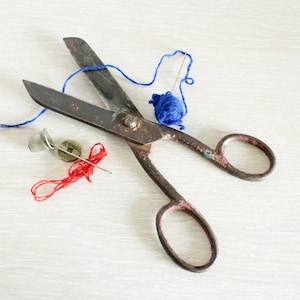May include: An overhead shot of vintage sewing tools. A pair of rusty scissors is surrounded by a blue yarn ball, a thimble, a wooden button, a needle with red thread, and loose blue thread.