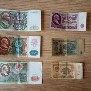 May include: A collection of vintage Soviet Union banknotes, featuring portraits and intricate designs. The bills display denominations of 5, 25, 50, 100, and 200 rubles, with various colors and details. The banknotes are arranged on a wooden surface.