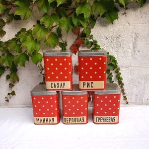 May include: A stack of five red metal storage containers with white polka dots. Each container has a silver lid and a label with Cyrillic text. The containers are arranged on a white surface, with green vines in the background.