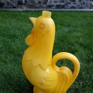 Könnte beinhalten: Eine hellgelbe, hühnerförmige Gießkanne. Die Kunststoffkanne hat einen Griff und einen Ausguss oben. Der Hahn hat einen detaillierten Kamm, Schnabel und Schwanzfedern. Die Kanne steht auf grünem Gras.