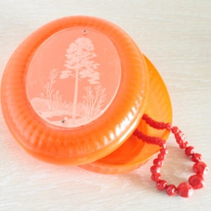 May include: An orange, round, vintage compact with a tree and landscape design etched on the lid. A red beaded necklace is draped over the compact. The compact is open, revealing a second orange section.