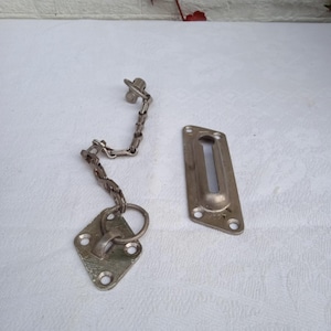 May include: A vintage silver-toned door chain and latch set. The set includes a diamond-shaped plate with a ring, a chain, a rectangular plate with a slot, and a small screw. The hardware is likely made of metal.