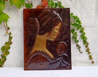 May include: A rectangular, brown relief sculpture of a woman holding a rose. The woman's profile is in gold, with her hair styled in a bun. The background is a darker brown with a quilted pattern. The sculpture is displayed against a white brick wall with green vines.