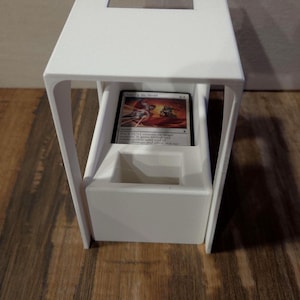 Puede incluir: Un soporte para cartas blanco impreso en 3D con un marco rectangular, una abertura superior y un compartimento central que contiene una carta coleccionable. La carta muestra ilustraciones y el texto Take Up the Shield.