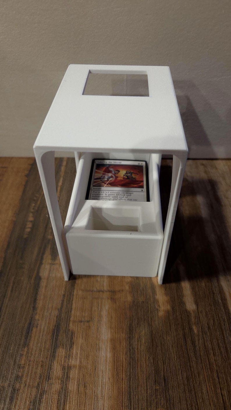 Puede incluir: Un soporte para cartas blanco impreso en 3D con un marco rectangular, una abertura superior y un compartimento central que contiene una carta coleccionable. La carta muestra ilustraciones y el texto Take Up the Shield.