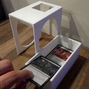 Puede incluir: Una caja para cartas impresa en 3D de color blanco con una tapa extra&iacute;ble que presenta recortes arqueados en los laterales. La caja est&aacute; abierta y muestra una pila de cartas coleccionables, con una carta parcialmente extra&iacute;da que muestra el texto Cruel Alliance y Teamwork 2.