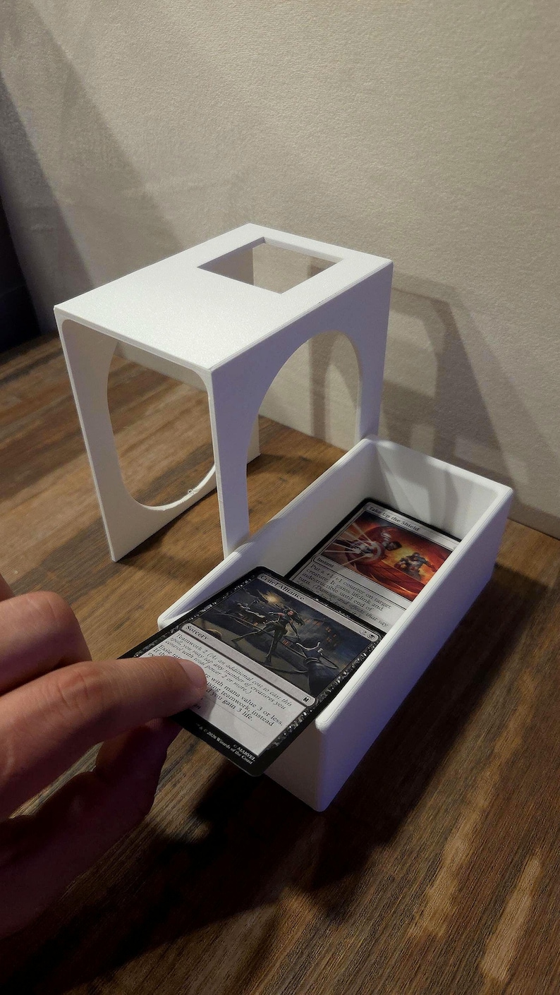 Puede incluir: Una caja para cartas impresa en 3D de color blanco con una tapa extra&iacute;ble que presenta recortes arqueados en los laterales. La caja est&aacute; abierta y muestra una pila de cartas coleccionables, con una carta parcialmente extra&iacute;da que muestra el texto Cruel Alliance y Teamwork 2.
