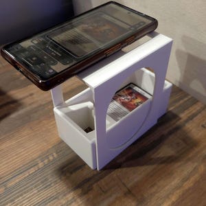 Puede incluir: Un soporte de pl&aacute;stico blanco con una superficie superior plana para un tel&eacute;fono inteligente y un compartimento inferior para guardar cartas coleccionables. El soporte presenta un recorte circular en el panel lateral y contiene una carta.