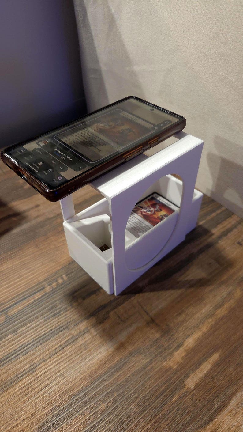Puede incluir: Un soporte de pl&aacute;stico blanco con una superficie superior plana para un tel&eacute;fono inteligente y un compartimento inferior para guardar cartas coleccionables. El soporte presenta un recorte circular en el panel lateral y contiene una carta.