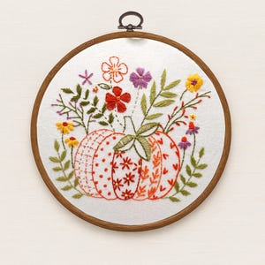 May include: Embroidered pumpkin hoop art featuring a detailed pumpkin design with orange, red, and white patterns. The pumpkin is surrounded by colorful embroidered flowers and green leaves. The artwork is set in a wooden hoop.