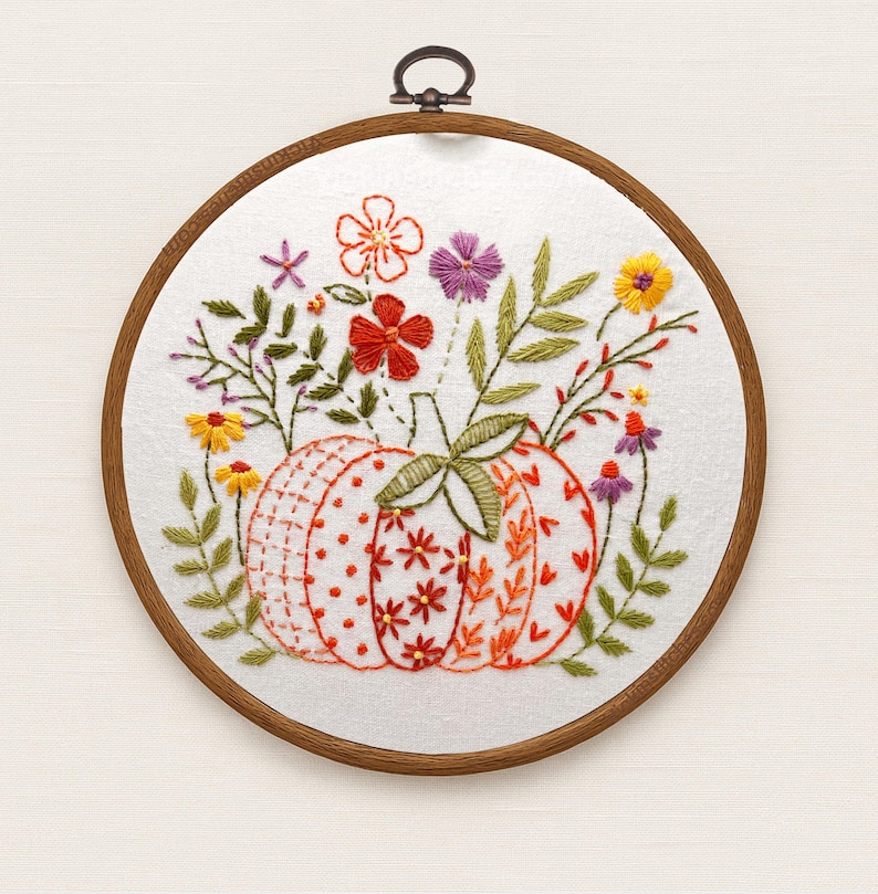 May include: Embroidered pumpkin hoop art featuring a detailed pumpkin design with orange, red, and white patterns. The pumpkin is surrounded by colorful embroidered flowers and green leaves. The artwork is set in a wooden hoop.