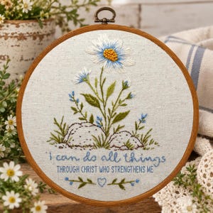May include: A circular wooden embroidery hoop framing a light-colored fabric with a hand-stitched design of a white daisy, green foliage, and two rocks. Below the floral motif, the text reads: I can do all things THROUGH CHRIST WHO STRENGTHENS ME.