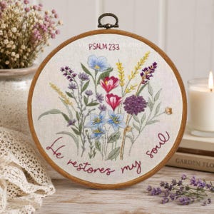 May include: A circular wooden embroidery hoop containing white fabric with a hand-stitched floral arrangement in shades of blue, pink, yellow, and purple. The text "PSALM 23:3" and "He restores my soul" is embroidered in a dark red cursive script.