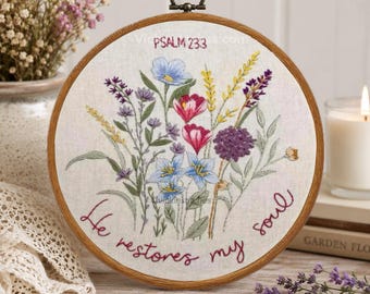 Puede incluir: Un bastidor de bordado circular de madera que contiene tela blanca con un arreglo floral cosido a mano en tonos azul, rosa, amarillo y morado. El texto "PSALM 23:3" y "He restores my soul" está bordado en una letra cursiva de color rojo oscuro.